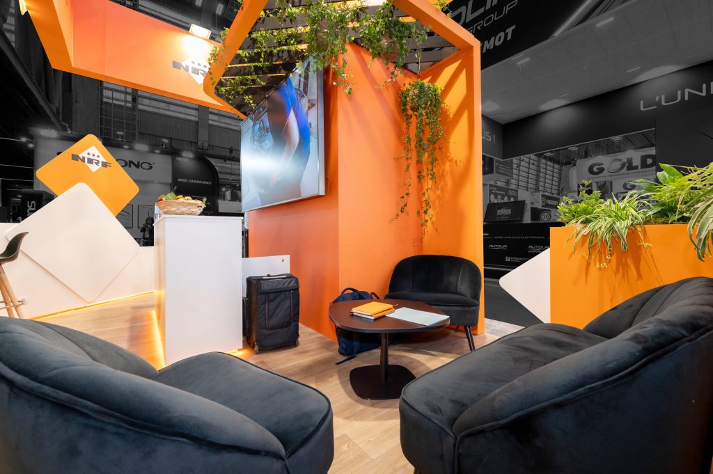 Vue de l'espace salon du stand sur-mesure aux couleurs orange et blanc réalisé par R2 Stand pour NRF sur le salon Equipauto avec podium produits, bar et écran géant
